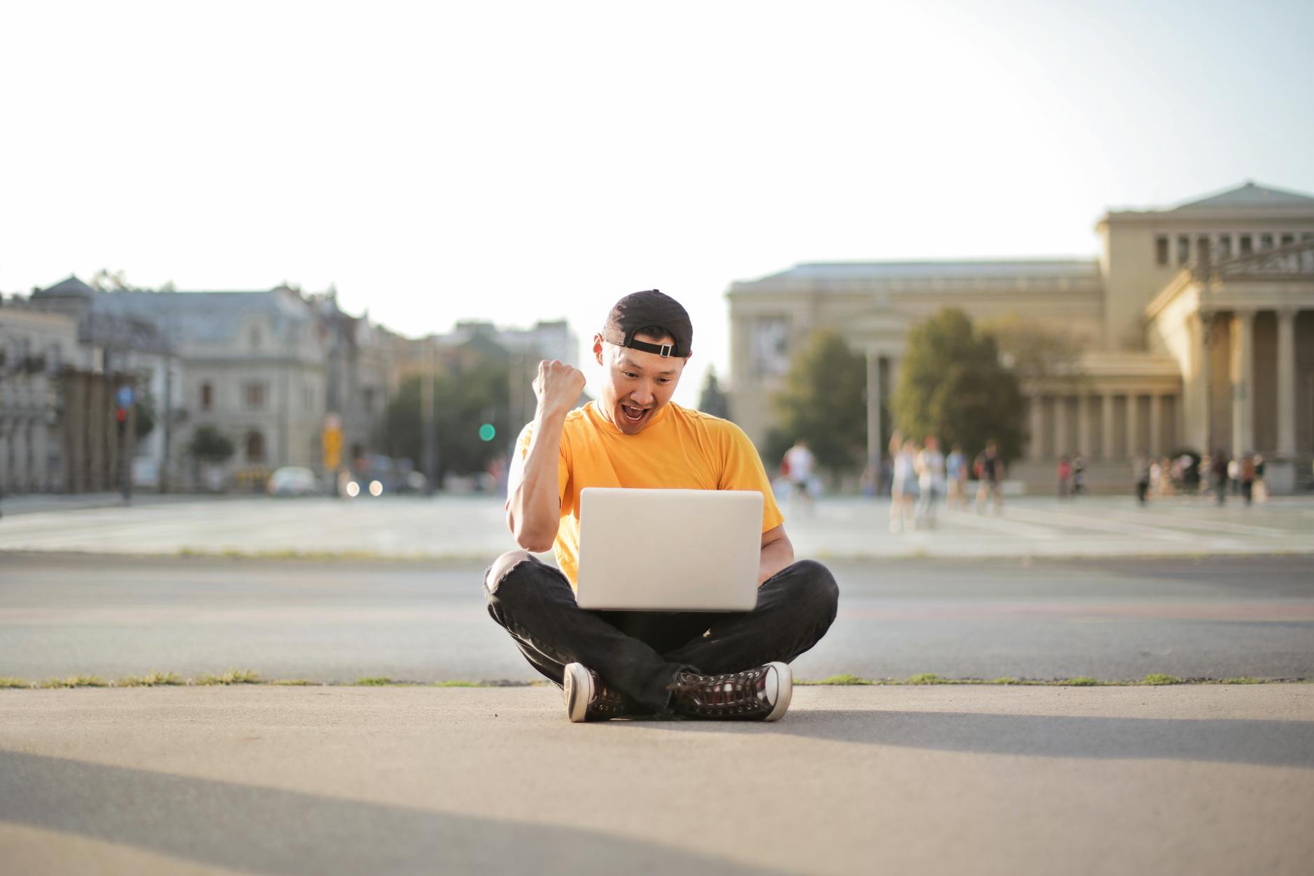 man in white t shirt and black pants sitting on the road using laptop computer